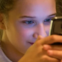 A teenage girl looks closely at her smartphone in a dimly lit room
