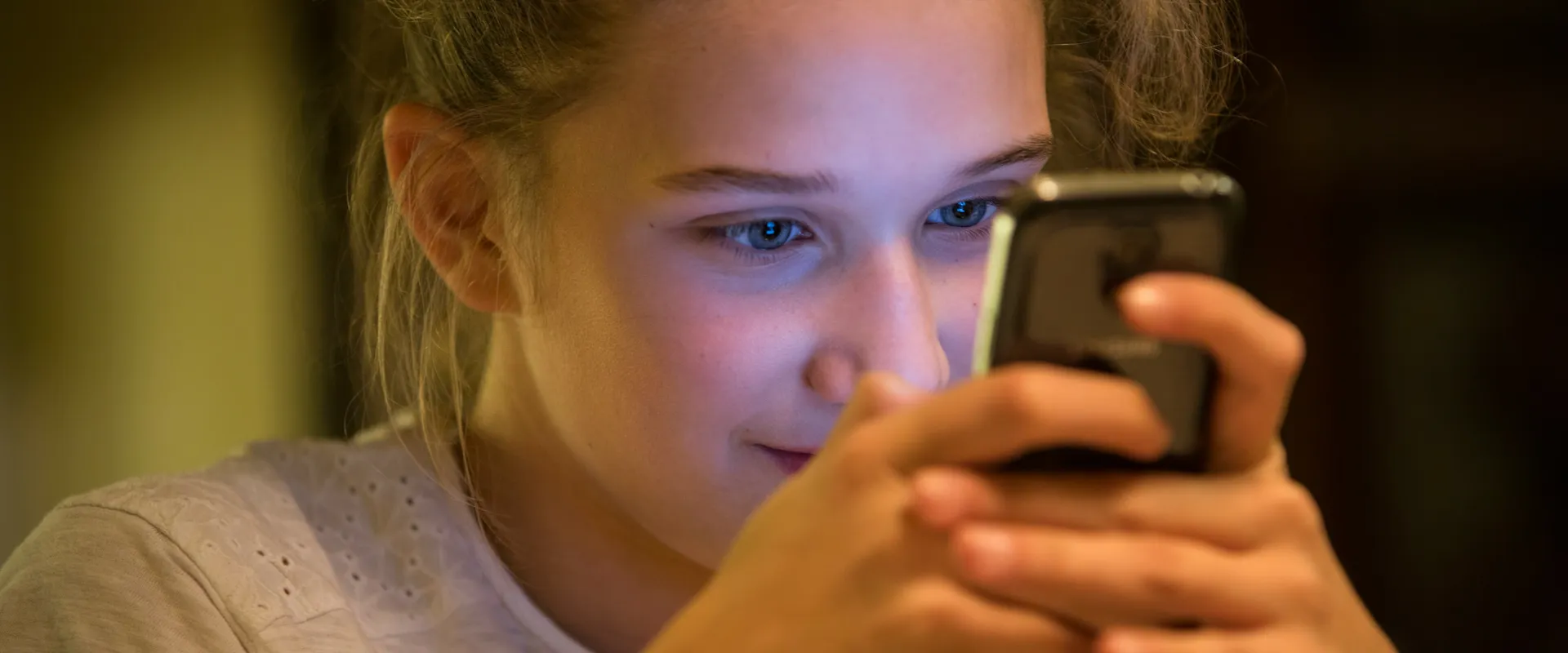 A teenage girl looks closely at her smartphone in a dimly lit room