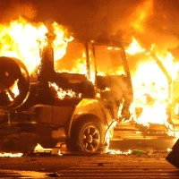 A silhouetted person carrying a stick walks past a burning SUV at night, with flames engulfing vehicle