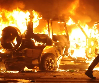 A silhouetted person carrying a stick walks past a burning SUV at night, with flames engulfing vehicle