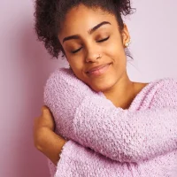 Young woman in a pink jumper hugging herself with her eyes closed and a contented smile