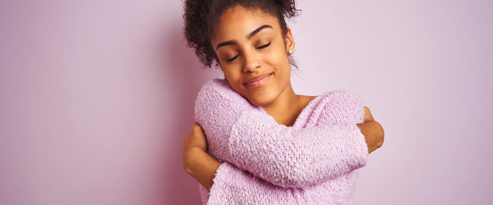 Young woman in a pink jumper hugging herself with her eyes closed and a contented smile